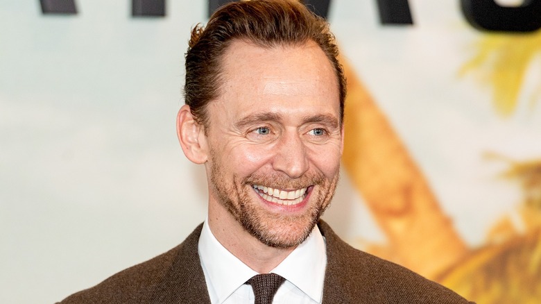 A man with coiffed brown hair flashes a big smile to the camera. He's wearing a brown jacket and tie.