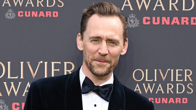 A man with slicked back brown hair and a short brown beard wears a black velvet tuxedo and a white shirt. He's stood against a black wall with the "OLIVIER AWARDS" logo on it in gold text. Underneath the logo is a crown logo with red text that reads "CUNARD"