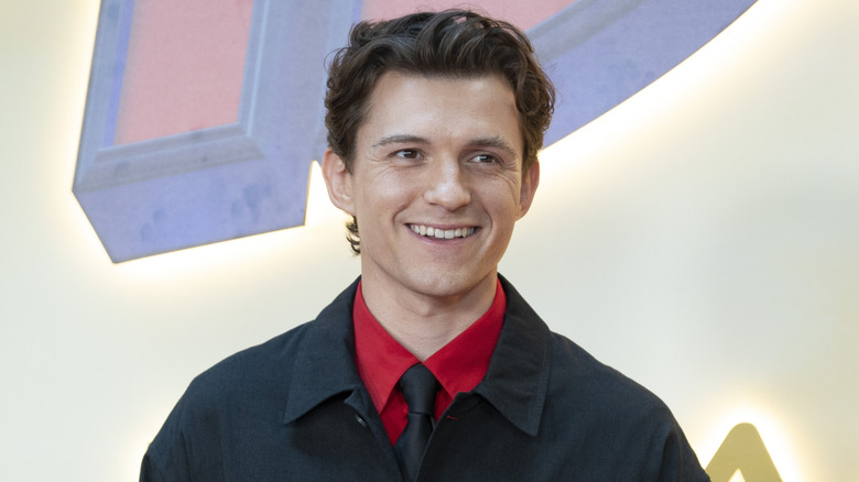 A young man with curly brown hair smiling while wearing a black button-up jacket with a red shirt and a black tie underneath.