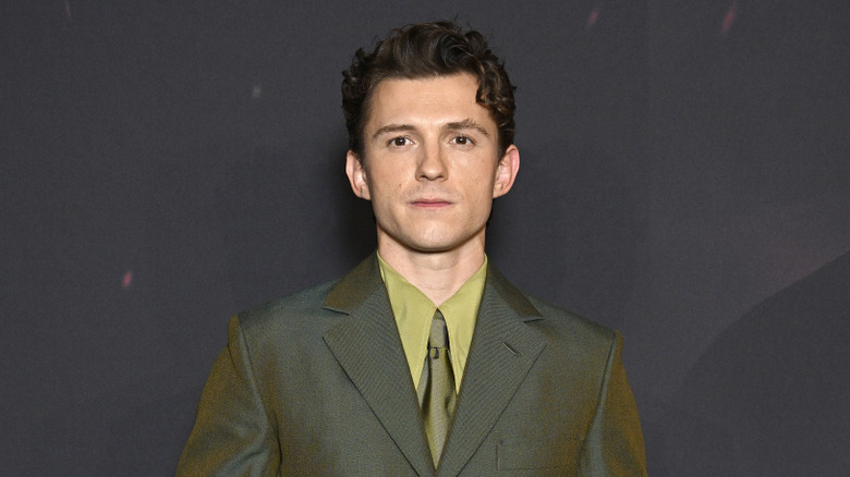 A young man with curly brown hair wearing a khaki green double breasted suit with a light green shirt and a khaki green tie.
