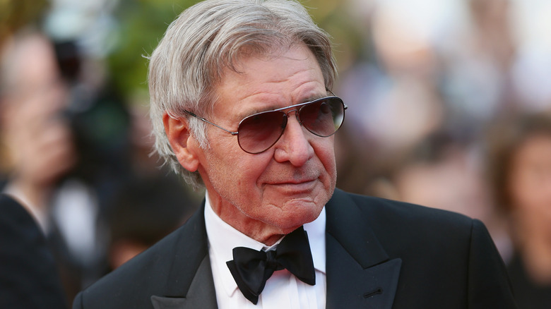 Harrison Ford with gray hair wearing a black tuxedo and aviator sunglasses.
