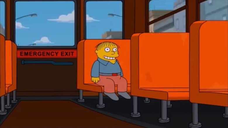 Family Guy/Simpsons geçişinden bir sahnede Ralph Wiggum, yüzünde bir gülümsemeyle okul otobüsünün arkasında oturuyor.