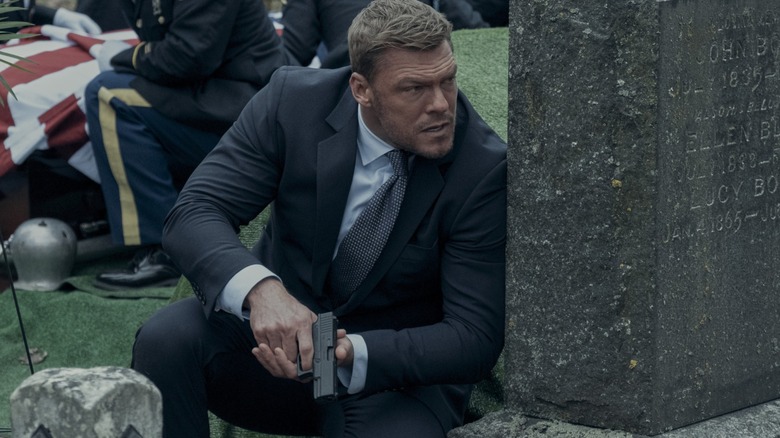 Jack Reacher holding a gun and ducking behind a grave