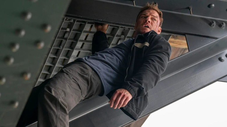 Jack Reacher hanging on the underside of a metal bridge one-handed