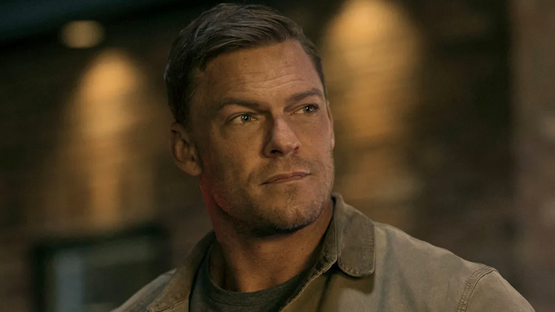 Alan Ritchson as Jack Reacher wearing a brown work jacket.