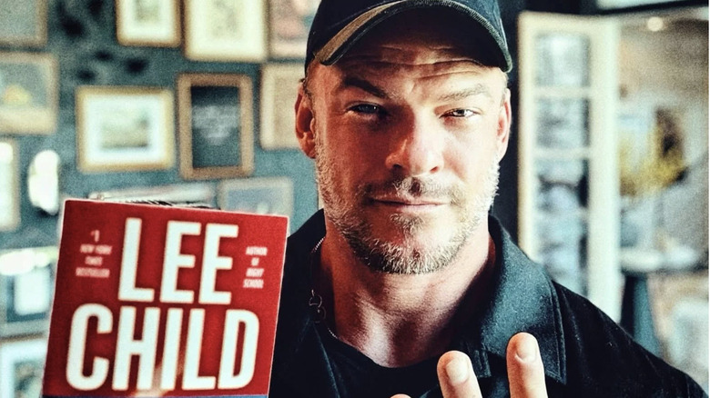 Alan Ritchson wearing a black baseball cap and black jacket while holding up a red book with the author's name "Lee Child" in white writing.