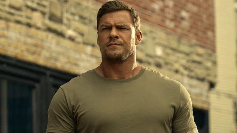 Alan Ritchson as Jack Reacher wearing an olive green t-shirt with brown hair, walking down a street with brick buildings behind him.