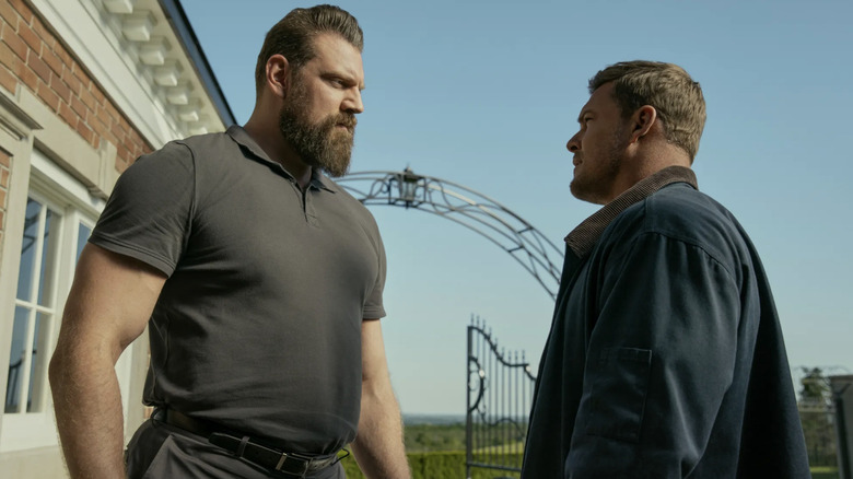Olivier Richters as Paulie with brown hair and a large brown beard, wearing a dark gray polo top, and Alan Ritchson as Jack Reacher looking up at Paulie, wearing a blue work jacket with a brown collar.