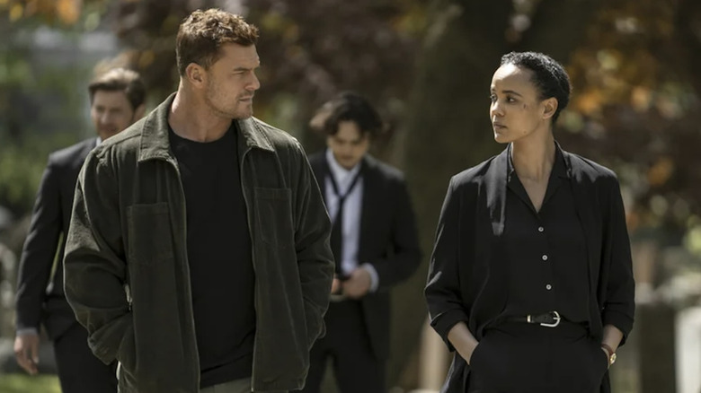 Alan Ritchson as Reacher wearing a green jacket and a black t-shirt, and Maria Sten as Frances Neagley wearing a black suit, walking through a graveyard with other funeral attendees behind them.