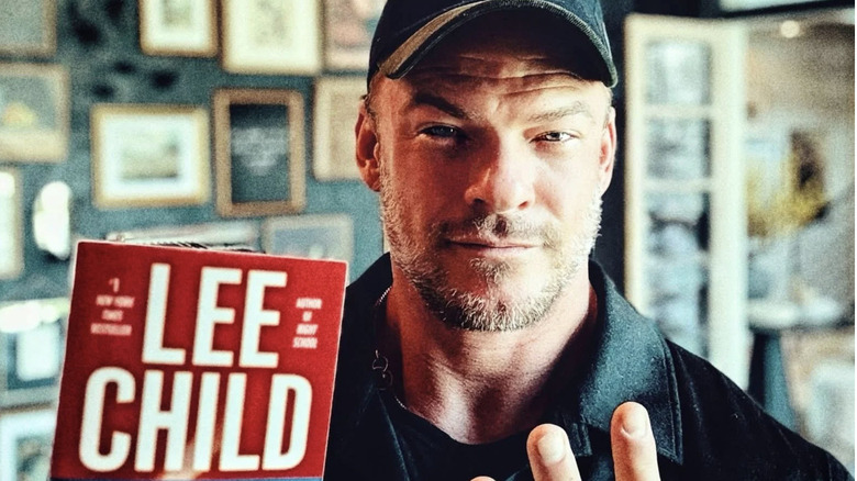 Alan Ritchson wearing a black baseball cap and black jacket while holding up a red book with the author's name "Lee Child" in white writing.