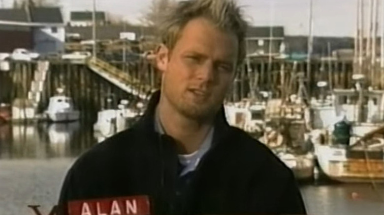 Blonde man standing in front of marina with banner underneath him reading Alan in Murder in Small Town X