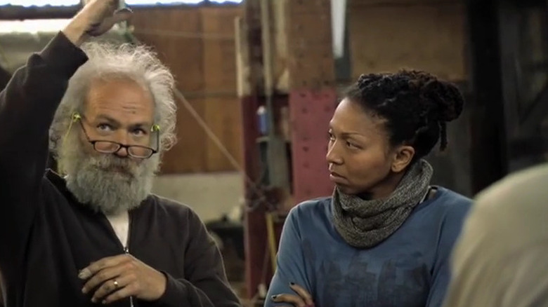 Man with white hair beard and glasses with hand up next to woman in blue shirt and scarf on The Colony