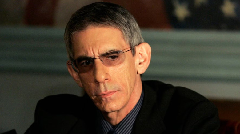 Detective Munch (Richard Belzer) sits at a desk wearing a black suit and glasses in 