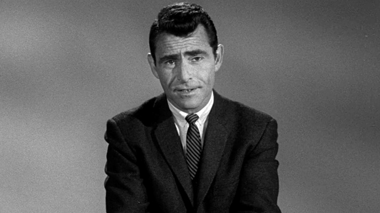 Rod Serling in a suit sitting down and looking at the camera in The Twilight Zone