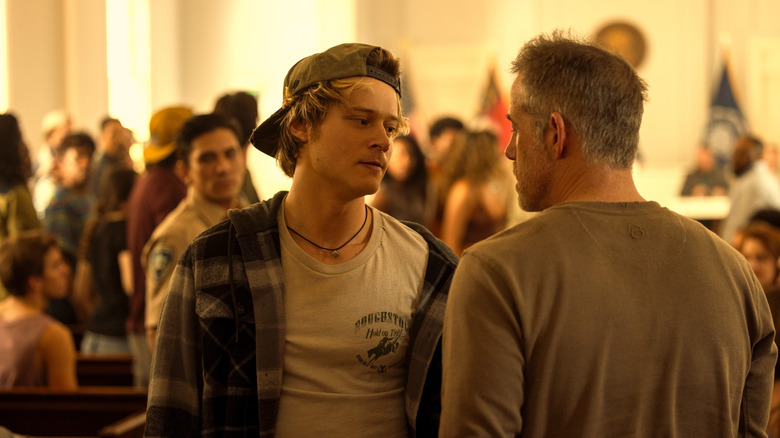 JJ wearing a backwards baseball cap and flannel shirt facing and speaking with Chandler Groff