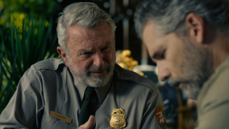 Solda Park Korucusu Şefi üniforması giyen gri saçlı ve sakallı Sam Neill, sağda ise haki gömlek giyen, griye çalan siyah saçlı Eric Bana var - Bana odak dışı.