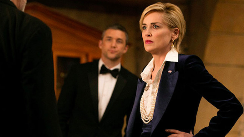 Sharon Stone as Natalie Maccabee with a serious expression in a navy blazer and pearls, looking at a man in the foreground, while Jeff Hephner as John Case stands behind her in a scene from "Agent X."