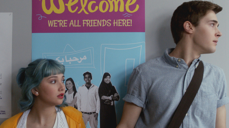 Amanda Arcuri as Lola Pacini and Eric Osborne as Miles Hollingsworth III in "Degrassi: Next Class," standing together in a school hallway in front of a colorful welcome poster.