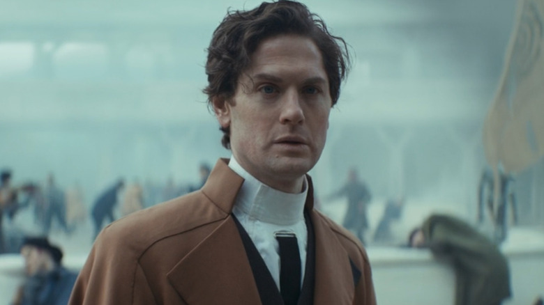 Kyle Soller as Syril Karn in "Andor," wearing a high-collared brown coat and white shirt with a panicked crowd behind him.