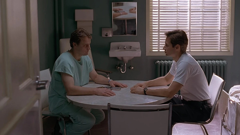 Robert Wisden as Robert Patrick Modell in turquoise hospital scrubs sitting at a table across from David Duchovny as Mulder with a revolver in between them in The X-Files