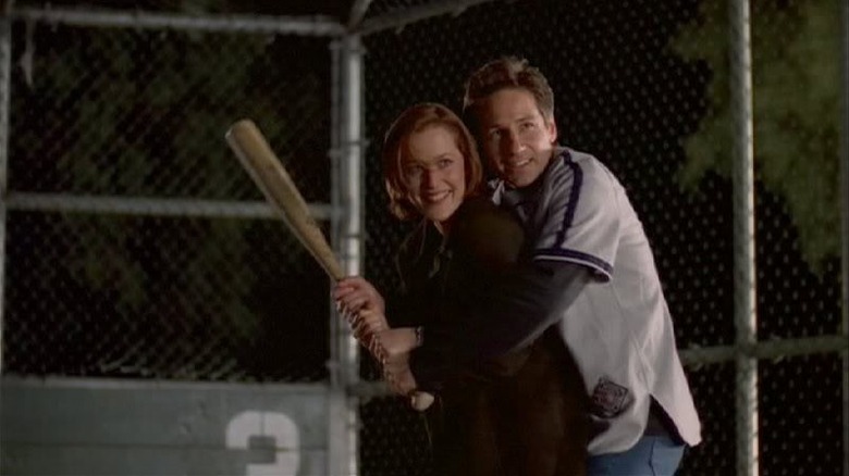 Gillian Anderson as Scully standing in front of David Duchovny as Mulder, both holding baseball bat and smiling in The X-Files