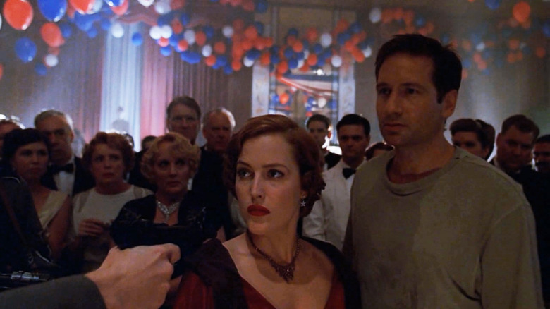 Gillian Anderson as Scully in period clothing and David Duchovny as Mulder in sweatshirt surrounded by people and balloons in The X-Files