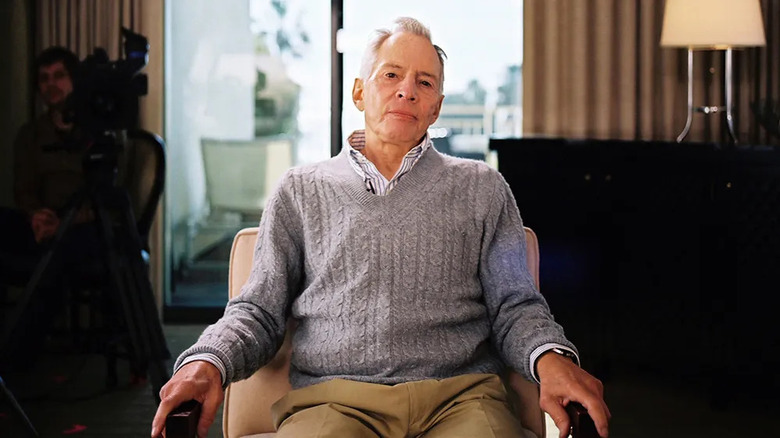 Robert Durst sits in a hotel room looking at the camera with a cameraman in the background in The Jinx