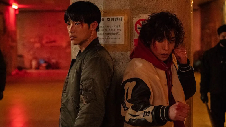 A battered Kim Geon-woo (Woo Do-hwan) and Hong Woo-jin (Lee Sang-yi) stand ready to fight by a concrete pillar in Bloodhounds