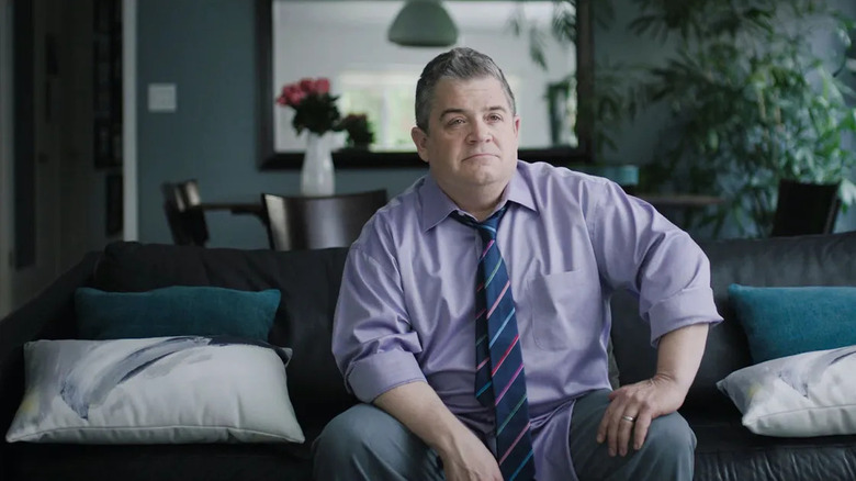 Patton Oswalt in purple shirt and tie sitting on couch in I'll Be Gone in the Dark