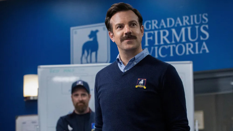 Ted Lasso (Jason Sudeikis) stands confidently in front of a white board and smiling Coach Beard (Brendan Hunt) in Ted Lasso