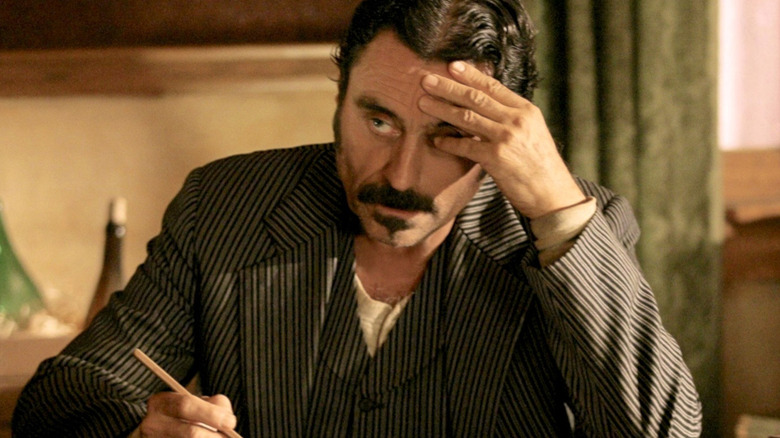 Al (Ian McShane) writing something looking stressed with his hand on his head in Deadwood