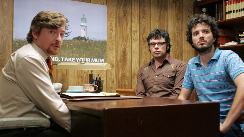 Murray (Rhys Darby), Jemaine Clement, and Bret McKenzie looking at something in the New Zealand tourism office in Flight of the Conchords