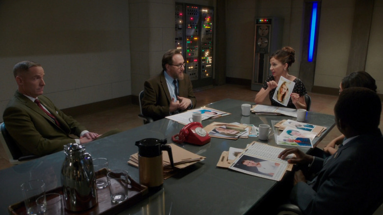 Val holds up a headshot of a Bad Place resident while sitting at a board room with demon boss Shawn and her coworkers Glenn, Babadjan and Gayle in "The Good Place Presents: The Selection"