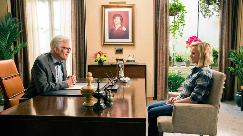 Michael sits behind a large wooden table with his hands crossed as he smiles at Eleanor sitting across from him in "The Good Place"