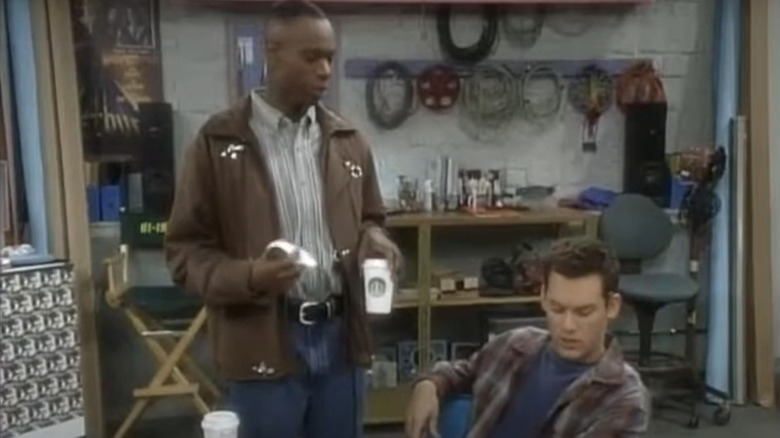 Dave stands next to John sitting at a desk in Buddies