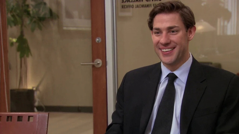 Jim Halpert (John Krasinski) sits smiling in an office in The Office