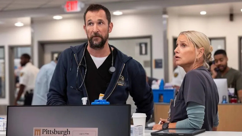 Dr. Robby Robinavitch rolünde Noah Wyle, siyah önlük üzerine lacivert bir sweatshirt ve boynunda bir stetoskop giyiyor, kameradan bakıyor ve Dana Evans rolünde Katherine LaNasa'nın yanında duruyor, gri bir önlük ve altında açık deniz mavisi uzun kollu bir gömlek giyiyor, dirseği The Pitt'teki tezgâhın üzerinde.