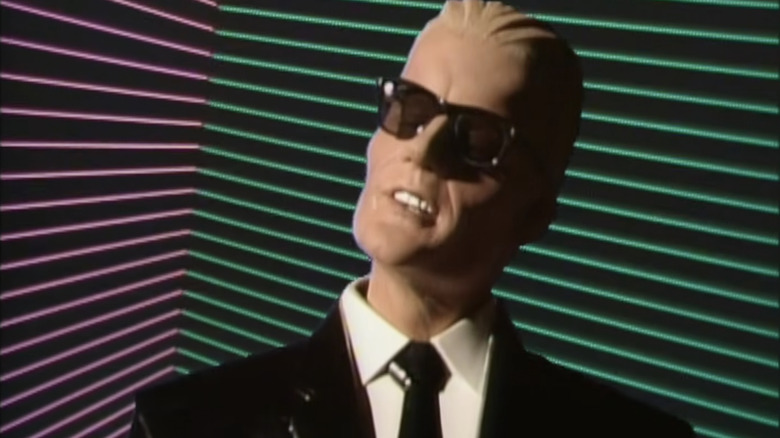 A "digital" man called Max Headroom with slicked back blond hair, black raybans and a plastic black suit and tie. He's stood in front of a neon-lined background that is green and pink.