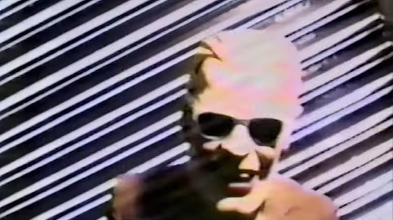 A fuzzy, distorted image of a person wearing a large "Max Headroom" mask with sunglasses. There is a silver lined background behind them.