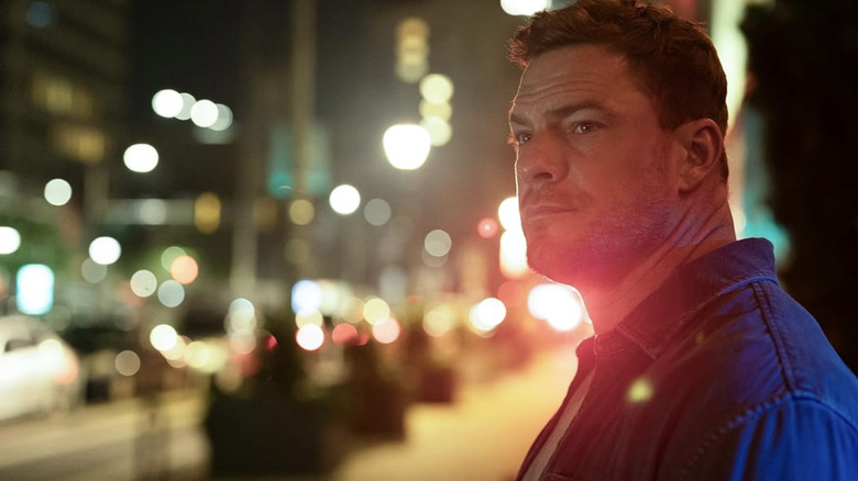 Jack Reacher looks to the side while lights from a city street glow behind him in Reacher