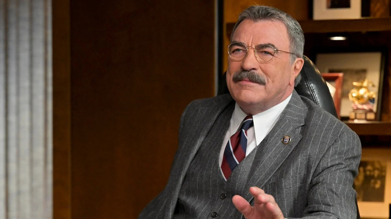 Tom Selleck, polis komiseri Frank Reagan rolünde gözlüklü, ince çizgili gri üç parçalı bir takım elbise, uzun yakalı bir gömlek ve Blue Bloods'da bir ofis koltuğunda otururken kamera dışındaki birine bakan beyaz şeritli kırmızı ve mavi bir kravat