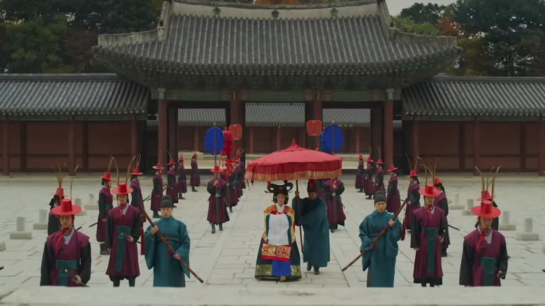 Korean courtyard with men wearing green and red and woman with red umbrella held over her in Kingdom