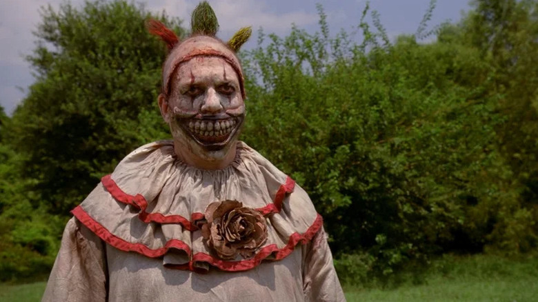 John Carroll Lynch's Twisty the Clown standing in front of lush greenery on American Horror Story: Freak Show