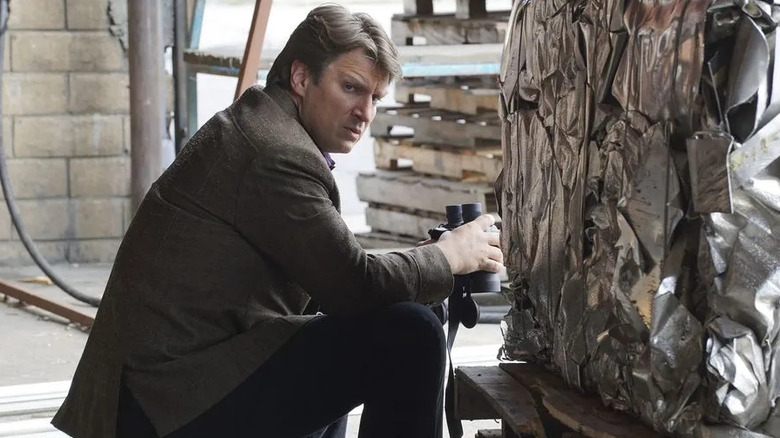 Richard Castle (Nathan Fillion) sits next to compressed metal while holding binoculars in Castle