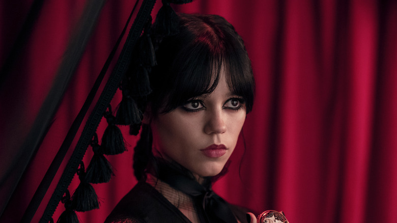 Jenna Ortega's Wednesday Addams dressed in black and standing against a red curtain on Wednesday