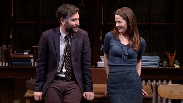 Aaron (Josh Radnor) and Joan (Elizabeth Reaser) sit against a wooden desk while smiling at each other in a stage production of The Babylon Line