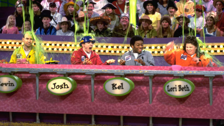 The ensemble of Figure It Out sits at a pink podium while being slimed in front of a studio audience