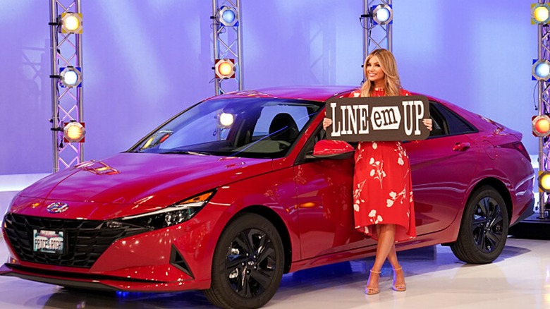 Amber Lancaster stands next to a new car while holding a sign that says "Line Em Up" on The Price is Right