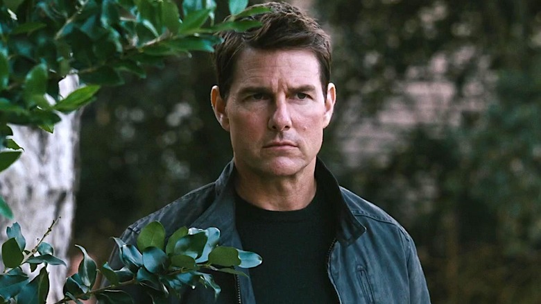 Jack Reacher in a black shirt and jacket standing among some leafy tree branches in Jack Reacher: Never Go Back