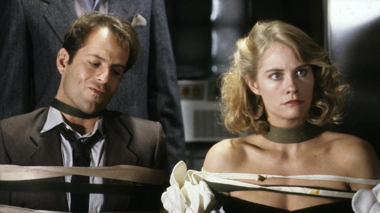 David (Bruce Willis) with an annoyed expression and Maddie (Cybill Shepherd) with wide eyes sit strapped to chairs by their bodies and necks in Moonlighting